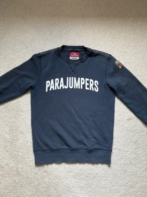 Mörkblå sweatshirt från Parajumpers - Mörkblå sweatshirt från Parajumpers med vit logotyptext framtill och patch på ena ärmen. Tröjan har rund hals, ribbade muddar och är tillverkad i mjuk bomullsfleece. Perfekt för en avslappnad och stilren look.