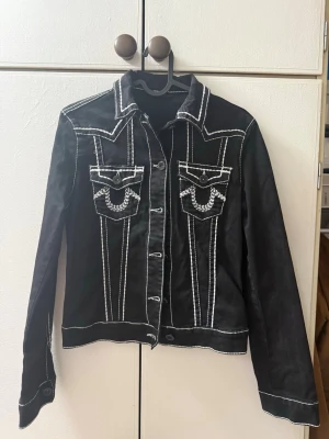 Black and white true religion jacket for women - Snygg svart jeansjacka med tydliga vita kontrastsömmar och två bröstfickor med lock och dekorativa detaljer. Klassisk krage och knappstängning framtill. Jackan har en cool streetstil och passar perfekt till jeans eller kjol.