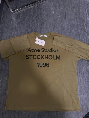 Mörk grön långärmad topp Acne Studios - Mörk grön långärmad topp från Acne Studios med svart texttryck 'STOCKHOLM 1996' framtill. Oversized fit och rund halsringning. Perfekt för dig som gillar streetwear och vill ha en statement piece i garderoben.