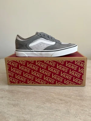 Gråa Vans sneakers - Snygga Vans sneakers i grå mocka och textil med vit sula och klassisk vit sidostrip. Skon har gråa skosnören, svart rand längs sulan och detaljer i ljusgrått. På hälen finns Vans-logga och siffrorna 66/99 broderat.