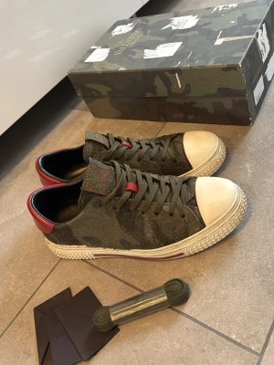 Camo sneakers från Valentino - Säljer ett par snygga sneakers från Valentino med camomönster i grönt och oliv, vita tåhättor och röda detaljer på häl och plös. Ej orginal sulor! Box, extra snören och valentino kort kommer med. Rickrigt schysta till sommaren. Skriv vid frågor!!