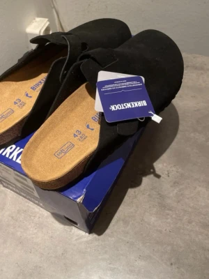 Svarta Birkenstock Boston tofflor - Svarta Birkenstock Boston tofflor i mocka med klassisk korksula och justerbart spänne. Sköna slip-in tofflor med anatomiskt utformad fotbädd som ger bra stöd. Perfekta för en avslappnad och stilren look.🏖️ realistad pga att jag inte gillade färgen men dom är väldigt fina annars plus dom var för stora