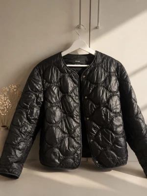 Svart quiltad pufferjacka från - Snygg svart pufferjacka  med quiltat mönster och rund halsringning. Jackan har tryckknappar framtill och en lätt glansig finish. Perfekt för kalla dagar när du vill hålla stilen.