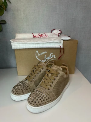 Beige sneakers från Christian Louboutin - Snygga beige sneakers från Christian Louboutin med nitar på tån och meshdetaljer på sidorna. Skorna har vita sulor, beige mocka och klassiska snörningar. Perfekta för dig som vill sticka ut med exklusiv design och edgy detaljer. 41 passar 42