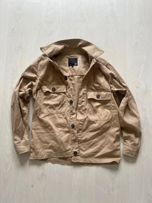 Beige overshirt från lager157 - Stilren beige overshirt från lager157 med två stora bröstfickor och knäppning framtill. Tillverkad i ett mjukt bomullstyg och har klassisk krage samt långa ärmar med knapp vid ärmslut. Perfekt att slänga över en t-shirt för en avslappnad look.