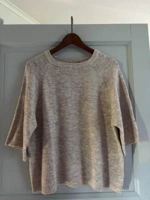 Beige stickad tröja med trekvartsärm - Mysig beige stickad tröja med trekvartsärmar och rund halsringning. Tröjan har en avslappnad passform och ribbstickade detaljer vid halsen. Perfekt för lager på lager och enkel att matcha till olika outfits.