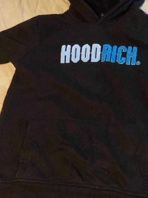 Svart HOODRICH hoodie med blå text - Snygg svart hoodie från HOODRICH med stor logga i vitt och blått på bröstet. Klassisk huva och magficka framtill. Perfekt för dig som gillar streetwear och vill ha en bekväm och stilren look. Mjuk bomullskänsla och avslappnad passform.