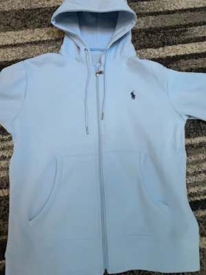 Ljusblå hoodie från Ralph Lauren - Snygg ljusblå hoodie från Ralph Lauren med dragkedja framtill och två fickor. Klassisk huva med dragsko och den ikoniska lilla loggan broderad på bröstet. Perfekt för en chill och avslappnad stil. Mjuk bomullskänsla och stilren design.