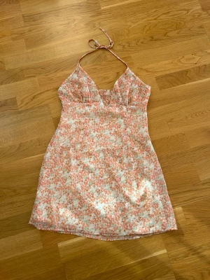  Ny Blommig kort sommarklänning - Söt kort klänning med blommigt mönster i rosa, vitt och ljusgrönt. Klänningen har halterneck med knyt i nacken och v-ringad urringning. Perfekt för varma dagar och har en lätt, luftig känsla.