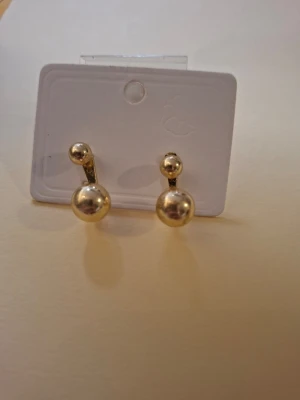 Guldtonade örhängen med pärldetalj - Snygga örhängen i guldtonad metall med en unik design där en liten pärla sitter ovanför en större guldfärgad kula. Perfekta för dig som gillar stilrena men ändå iögonfallande accessoarer.