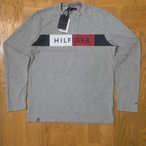 Grå långärmad topp Tommy Hilfiger - Snygg grå långärmad topp från Tommy Hilfiger med logga och färgblock i vitt, marinblått och rött över bröstet. Tillverkad i mjuk bomull och har en klassisk passform. Perfekt för en clean och stilren look.