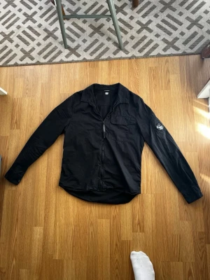 Svart overshirt från C.P. Company - Riktigt snygg overshirt/jacka passar perfekt nu när det börjar bli lite varmare och tänkte själv ha den men den passar inte längre. Den är äkta som du ser på bilden och om du är osäker på om den är äkta så scanna koden och skriv in CLG koden så ser du själv