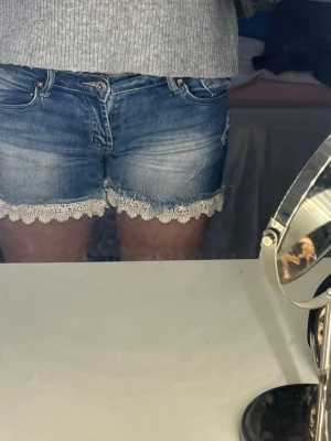 Blå jeansshorts med spetskant - Säljer ett par blå jeansshorts med snygg spetskant i vitt längst ner. Lågmidjade och perfekt längt! Stl xs/S