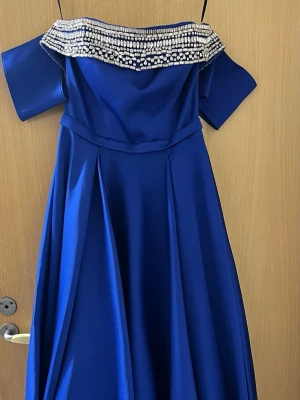 Blå långklänning med pärldetaljer - Elegant blå långklänning med offshoulder-design och bred pärlbesatt kant upptill. Klänningen har en utsvängd kjol och är tillverkad i ett glansigt material som ger en lyxig känsla. Perfekt för bal eller festliga tillfällen.
