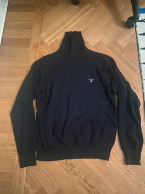 Svart polotröja från Gant - Klassisk svart stickad polotröja från Gant med diskret logga och sköld på bröstet. Tröjan har lång ärm, ribbade muddar och hög polokrage. Perfekt för dig som gillar stilrena och tidlösa plagg.