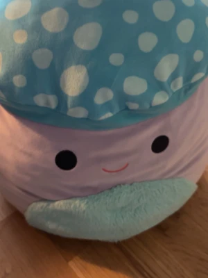 Stor Squishmallow manet - Säljer en stor Squishmallow i form av en söt manet med ljuslila kropp, blått huvud med vita prickar och ett leende ansikte. Tillverkad i supermjuk plysch, perfekt som kudde eller mysig inredningsdetalj.