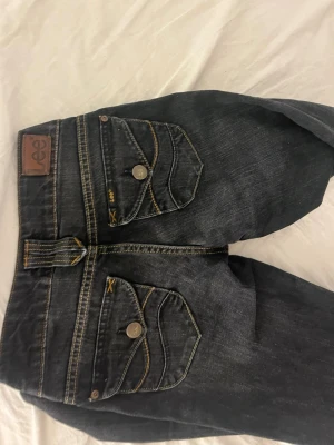 Mörk blåa lågmidjade jeans - Säljer ett par mörkblåa  jeans från Lee med snygga kontrastsömmar och breda bälteshällor. Jeansen har klassisk femficksdesign och raka ben. Materialet är denim i bomull med en lätt tvättad look. Perfekta för en avslappnad och trendig stil.