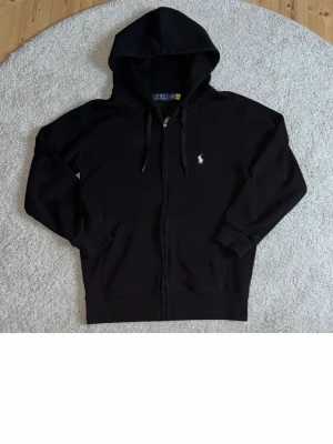 Svart hoodie från Polo Ralph Lauren - Svart hoodie med dragkedja från Polo Ralph Lauren. Klassisk design med huva och dragsnören, två fickor framtill och den ikoniska vita logotypen broderad på bröstet. Tillverkad i mjukt material som känns skönt mot huden.