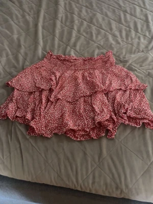 Röd volangkjol med blommigt mönster - Supersöt kort kjol med volanger i rött och vitt blommigt mönster. Kjolen har resår i midjan och flera lager volanger som ger en lekfull look. Perfekt för varma dagar och enkel att matcha med en enkel topp.