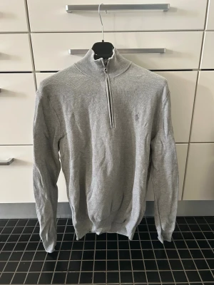 Grå stickad halvzip från Ralph Lauren - Snygg grå stickad tröja från Ralph Lauren med halv dragkedja och hög krage. Tröjan har diskret logga på bröstet och är tillverkad i mjukt material. Perfekt till lager på lager och enkel att matcha med jeans eller chinos. Tröjan har knappt använts. 