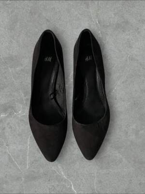 Svarta pumps från H&M med smal klack - Stilrena svarta pumps från H&M med spetsig tå och smal klack. Skorna har en klassisk siluett och är tillverkade i ett material som liknar mocka. Perfekta för att lyfta vilken outfit som helst. Små i storlek 
