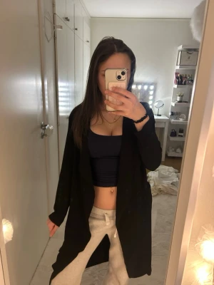 Svart lång trenchcoat med skärp - Stilren svart trenchcoat med öppen front och löst fall. Jackan har ett tillhörande skärp i midjan och långa ärmar. Perfekt för lager på lager och ger en snygg siluett. Materialet ser ut att vara mjukt och lätt, vilket gör den enkel att bära.