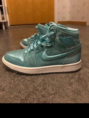 Turkosa Air Jordan 1 High sneakers - Säljer ett par Air Jordan 1 High sneakers i en unik turkos färg. Skorna har en blank satinliknande finish, klassisk Nike Swoosh på sidan och Air Jordan-logga vid ankeln. Vita sulor och matchande turkosa skosnören.