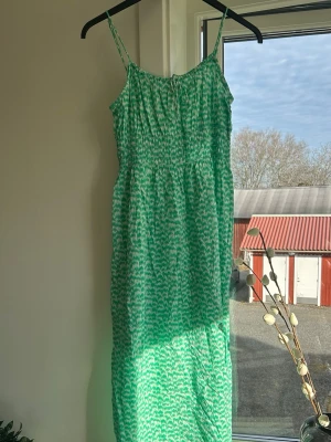 Grön mönstrad långklänning - Säljer en grön långklänning med smala axelband och ett vitt abstrakt mönster. Klänningen har smock upptill för en bekväm passform och är tillverkad i ett lätt material som passar perfekt till varma dagar💚