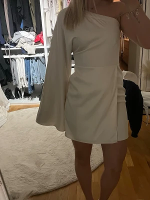 Vit oneshoulder kortklänning - Stilren vit kortklänning med oneshoulder-design och en lång, vid ärm. Klänningen har en figurnära passform och är tillverkad i ett mjukt, slätt material som ger en elegant look. Perfekt för dig som vill sticka ut med en unik siluett.