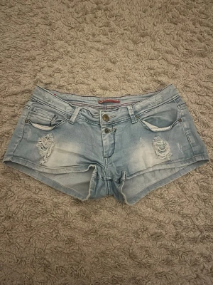 Ljusblå denimshorts med slitningar - Säljer ett par ljusblå denimshorts med slitna detaljer och spets bakom de (man kan se på 4de bilden) . Shortsen har fickor, dubbla knappar och dragkedja framtill. Perfekta för sommaren och ger en avslappnad streetstyle-vibe. Jeansshortsen är köpta utomlands och jag skulle säga att jeansshortsen är stora i storleken, skriv ifall frågor kommer upp!🩷🩷