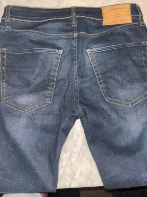 Blå jeans från Jack & Jones - Säljer ett par blå jeans från Jack & Jones med klassisk femficksdesign och snygga slitningar. Jeansen har kontrastsömmar och en läderpatch bak i midjan. Materialet är denim och passformen är rak. Perfekt för dig som gillar en avslappnad stil.