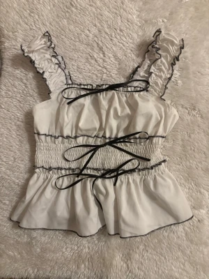 Vit volangtop med svarta detaljer - Supersöt vit linnetopp med volangkantade axelband och svarta kontrastsömmar. Toppen har smock i midjan och dekorativa svarta rosetter framtill. Perfekt för en trendig och feminin look.