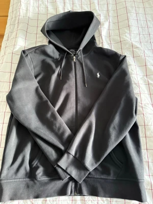 Svart zip från Ralph Lauren - Snygg svart hoodie från Polo Ralph Lauren med dragkedja framtill och klassisk huva med dragsko. Ikoniska vita loggan broderad på bröstet. Tillverkad i mjukt bomullsmaterial och har två fickor framtill. Perfekt för en chill och stilren look. I extremt bra skick och inga defekter vad jag kan se. Hör av er om ni har några frågor eller bud! 