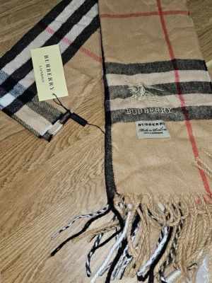 Beige rutig halsduk från Burberry Cashmere - Klassisk beige halsduk från Burberry i 100% kashmir med det ikoniska rutmönstret i svart, vitt och rött. Halsduken har fransar i ändarna och broderad Burberry-logga. Perfekt accessoar för att lyfta din outfit.