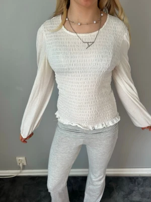 Vit långärmad smockad topp - Säljer en vit långärmad topp med smockad kropp och lätt volangkant nedtill. Toppen har rund halsringning och är tillverkad i ett mjukt, stretchigt material som sitter snyggt på kroppen. Perfekt att styla med jeans eller kjol.