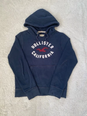 Mörkblå hoodie från Hollister - Mörkblå hoodie från Hollister med stor huva och magficka. Framtill står det 'Hollister California' i vitt och rött broderat märke. Klassisk passform och mjukt material, perfekt för chill dagar. Snygg logga på bröstet och ribbade muddar vid ärmar och nederkant.