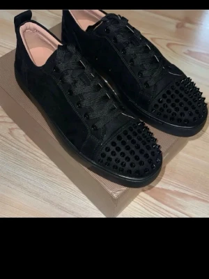 Svarta Louboutin sneakers med nitar - Säljer ett par svarta sneakers från Christian Louboutin med mattsvarta nitar på tån och sidorna. Skorna har svart mocka, svarta skosnören och den ikoniska röda sulan. Snygg och edgy design som sticker ut.