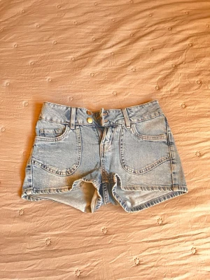 Ljusblå denimshorts med broderi - Säljer ett par ljusblå denimshorts med hög midja och snygga broderade detaljer på bakfickorna. Klassisk femficksmodell med knapp och dragkedja framtill. Perfekta för sommaren och ger en avslappnad vibe.