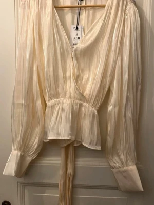 Krämfärgad omlottblus från Gina Tricot - Säljer en elegant krämfärgad omlottblus från Gina Tricot. Blusen har långa, lätt puffiga ärmar med breda manschetter, djup v-ringning och knytband i midjan. Materialet är glansigt och ger ett lyxigt intryck. Perfekt för dig som gillar trendiga detaljer.