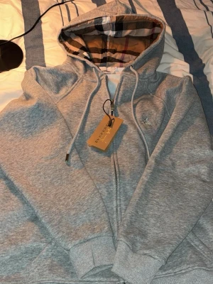 Grå zip hoodie från Burberry - Snygg grå zip hoodie från Burberry med klassiskt rutigt foder i huvan och dragsko. Broderad logga på bröstet och ribbade muddar vid ärmar och nederkant. Perfekt för dig som gillar stilrena och exklusiva plagg. Tillverkad i mjuk bomullsblandning.