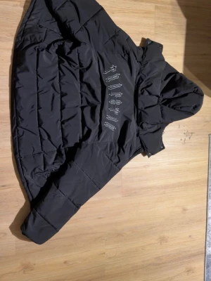 Svart pufferjacka från Trapstar - Säljer en svart pufferjacka från Trapstar med stor broderad logga på ryggen. Jackan har huva och quiltad design som ger en riktigt fet streetwear-look. Perfekt för kalla dagar och riktigt snygg till alla outfits.