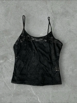 Svart spetsig linnetopp - Säljer en svart, ärmlös linnetopp med tunna axelband och genombruten spets. Toppen har en croppad passform och är perfekt för att styla lager på lager. Enkel men ändå edgy look.