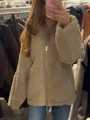 Beige teddyjacka med dragkedja - Mysig beige teddyjacka med dragkedja framtill och oversized passform. Jackan har stora ärmar och mjukt, fluffigt material som ger en avslappnad look. Perfekt för att hålla värmen under kyliga dagar och enkel att matcha med jeans eller leggings.