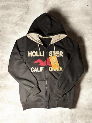 Svart hoodie från Hollister med tryck - Snygg svart hoodie från Hollister med dragkedja, beige insida på huvan och kontrasterande snören. Framsidan har stort tryck med texten 'Hollister California' och en röd logga. Perfekt för en avslappnad och trendig stil.