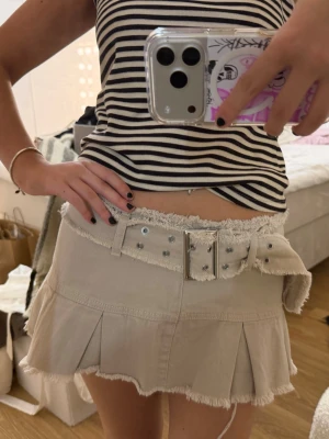 Beige kortkjol med fransad kant och bälte - Trendig beige kortkjol med fransade kanter och matchande bälte med nitar. Kjolen har plisserade detaljer nedtill och ger en cool, edgy vibe. Perfekt för dig som vill sticka ut med en unik och snygg look.