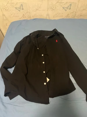 Svart skjorta från Ralph Lauren - Klassisk svart skjorta från Ralph Lauren med röd broderad logga på bröstet. Skjortan har lång ärm, knappar framtill och är i bomull. Perfekt för en clean och stilren look.