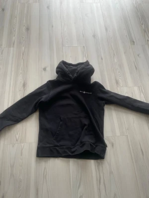 Svart hoodie från Sail Racing - Svart hoodie från Sail Racing med vit logga på bröstet. Klassisk modell med huva och dragsko samt stor magficka framtill. Tillverkad i mjukt material som känns skönt mot huden. Perfekt för en avslappnad och sportig stil.