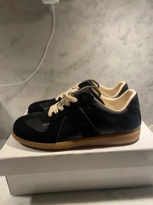 Svarta sneakers från Maison Margiela - Svarta sneakers från Maison Margiela med beige sula och vita skosnören. Ovandelen är i en mix av mocka och läder, med klassisk tysk träningsdesign och diskret logga på plösen. Insidan är ljusbeige och sulan har en retro gummifinish.