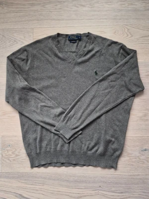 Ralph Lauren V-Neck Sweater - Grå Ralph Lauren V-Neck Sweater med grön emblem. Storlek M. Har ett väldigt litet hål på armen som knappt syns (sista bilden)  Alexbredd:52cm Längd:67cm  Lägg ett bud om du är intresserad :) Skickar inom 24 timmar!