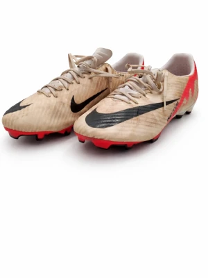Nike Mercurial air zoom fotbollsskor beige/röd - Säljer ett par Nike Mercurial fotbollsskor i beige med svarta swoosh-loggor och röda detaljer på hälen. Skorna har snörning och dobbar för spel på gräs. Tillverkade i syntetmaterial för lätt känsla och bra grepp på planen.